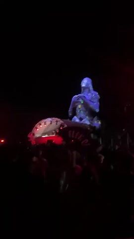 The video captures a nighttime outdoor event, likely a music festival or concert. A large, illuminated sculpture of a human figure is projected with vibrant, shifting colors, standing tall against the dark sky. Below it, a crowd of people is silhouetted against red and blue stage lights. In the foreground, a person holds up a large red fan with the word "FUCK OFF" written in white letters. Another person in the crowd is wearing a white t-shirt with the word "HAWAII" on it. A stage with a DJ booth and lighting equipment is visible behind the crowd. The overall atmosphere appears energetic and celebratory.