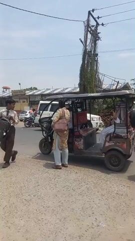 The video shows a street scene in India. In the first shot, an auto-rickshaw is stopped on a paved road. Two people are walking away from the auto-rickshaw, one of them wearing a light-colored hijab. A person is sitting in the auto-rickshaw. Other vehicles are nearby. Behind the vehicles is a utility pole with power lines, and trees and buildings are further back.  

The next shots show a motorcycle with two riders passing the camera. The camera is then moved to show storefronts along the roadside. Some of the shops are closed. There are also several white SUVs parked along the road. Another motorcycle drives by. A trailer is parked at the side of the road in the final shot.  The sky is light blue with some clouds.