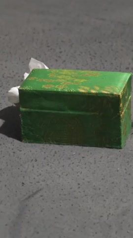 The video displays a closed, rectangular, green box with gold patterns. The box is positioned on a grey, textured surface. A small cluster of white material, possibly tissue paper, is visible peeking out from the left side of the box. The lighting appears dim, casting a shadow to the right of the box. The camera remains stationary throughout the short clip.