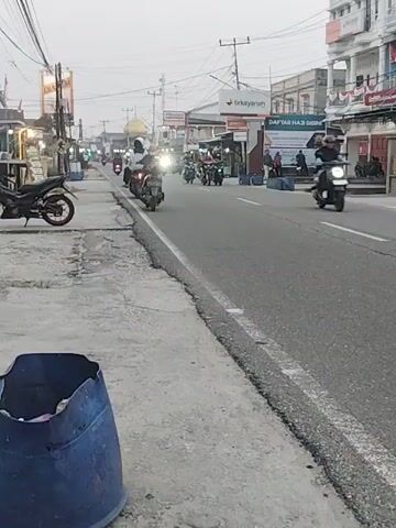 A street scene in what appears to be Southeast Asia is captured in this video. The time of day seems to be either early morning or late afternoon, as the lighting is soft and diffused. The road is paved and has white lane markings. Several motorcycles are visible on the road, some moving and some stationary. On the left side of the road, there is a sidewalk with a blue trash bin in the foreground. Behind the trash bin, a black motorcycle is parked. Further down the sidewalk, a few more motorcycles are parked or being tended to by people. On the right side of the road, there are buildings that appear to be shops or businesses, with awnings and signs. Some people are visible walking or standing near the buildings. One building has a large sign that reads "BRYKAYARLAH". Another sign in the distance indicates a "DAFTAR HAJI DOMPET" which translates to "Hajj Savings Registration". The overall atmosphere suggests a moderately busy urban or semi-urban environment.