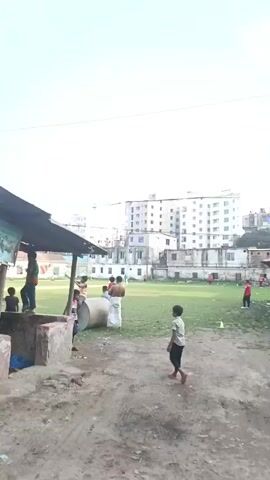 A medium-shot, slightly angled perspective video shows a group of people playing cricket on a grassy field surrounded by urban development. Several children are standing near a concrete structure and a large metal cylinder in the foreground. One child walks towards the field. Beyond the immediate area, several buildings of varying heights are visible, some under construction, set against a bright sky. Power lines run across the top of the frame. A person without a shirt, wrapped in a white cloth, stands near the cylinder with other people. On the cricket field, several players are actively engaged in the game. In the foreground near the camera, there are some cardboard boxes.
