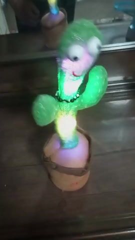 On a wooden surface, a dancing cactus toy sways and emits light while a person in the background observes. The toy, green and glittery, with large, cartoonish eyes, is in a brown fabric pot. It is equipped with a black beaded necklace. The lighting cycles between rainbow-like colors and green. The person stands out of focus, a reflection from a mirror on a dresser or cabinet.