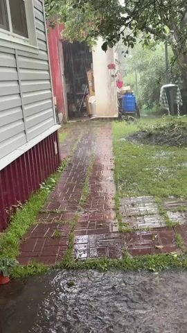 The video shows a torrential downpour in a rural backyard. Heavy rain is visible falling, creating puddles and streams on a brick pathway and the surrounding grass. A large, grey siding building is on the left, with a red-trimmed base. To the right of the pathway is a red garage-like structure with an open bay, revealing shelves and various items stored inside. Beyond the garage, a blue barrel and a white object covered with a checkered cloth are visible. The overall environment suggests a garden or storage area during a severe rainstorm.