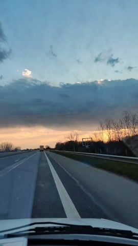 The video shows a view from the inside of a car driving on a highway during a sunset. The sky is a mix of light blue and dark grey clouds, with the setting sun casting a warm yellow glow. On the right side of the road is a metal guardrail, beyond which lies a strip of trees and vegetation. A large truck can be seen in the distance on the left side of the road. The dashboard of the car is visible at the bottom of the frame.  There is music playing.