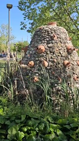 The camera captures a slightly low-angle, medium shot of a peculiar cone-shaped structure in an outdoor setting. The structure is composed of rocks and numerous terracotta pots of varying sizes embedded within the rockwork. On top of the cone, a larger terracotta pot serves as the apex. The base of the structure is surrounded by a variety of plants, including tall, green reeds, broad, leafy ground cover, and some cattails.

A tall metal lamppost stands to the left of the structure, reaching upwards against a clear, bright blue sky. In the background, there are trees with green foliage and some buildings that appear to be residential. A street with cars is also visible in the distance. The lighting indicates a sunny day, with shadows cast by the plants and the structure itself. Overall, the scene has a natural, park-like feel, with the unusual structure serving as a focal point.