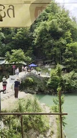 The video shows a scenic view of a river with a stone bridge spanning across it. Several people are walking on the bridge, and some are standing on the riverbank. The surrounding area is lush with green trees and vegetation. The water in the river is a clear, turquoise color. There are small buildings and structures visible on the riverbanks, suggesting a tourist or recreational area. One person is seen jumping from the bridge into the water, creating a splash. The overall atmosphere appears to be one of leisure and natural beauty.