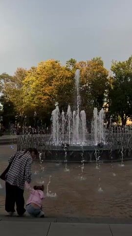 A woman and a child are in a park, near a fountain. The woman is wearing a plaid jacket and black pants. The child is wearing a pink hoodie and blue jeans. They are both looking at the fountain, which has water jets shooting up into the air. Some of the water jets are illuminated with pink and blue lights. Trees with yellow and green leaves are in the background. The sky is overcast.