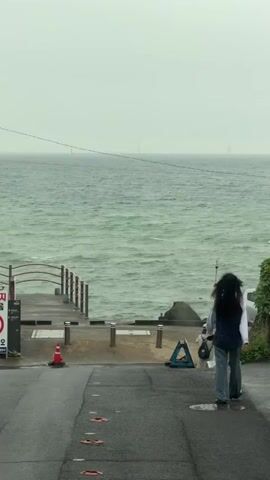 The video shows a view of the ocean on a cloudy day. In the foreground, a woman with long dark hair walks away from the camera down a wet paved path. She is wearing a backpack. To her left, there is a wooden pier with a red traffic cone on it. Further in the background, the ocean is choppy, and the sky is overcast. There are distant structures visible on the horizon, possibly wind turbines or lighthouses.
