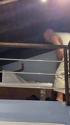 The video shows a person on a raised platform with a bed. Another person is leaning over the railing of the platform, looking down. The person on the platform is partially covered by a white sheet and is sitting up, reaching out a hand. The person leaning over the railing is wearing a white tank top and khaki shorts. The environment appears to be an attic or loft space with wooden beams on the ceiling. There are two lights visible on the ceiling. A plastic water bottle and some dark footwear are visible on the floor below the platform. The video is shaky and appears to be handheld.