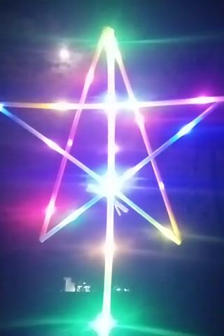 The video begins by showing a star-shaped object illuminated with colorful lights in the dark. The object appears to be made of thin, translucent rods connected to form a star. The lights are a mix of blue, pink, yellow, and green. There is a bright light source in the center of the star, and the entire object is glowing. The background is dark, suggesting it is nighttime. As the video progresses, the lights on the star fade out, and the object becomes almost invisible against the dark background. A single light source, possibly a street lamp or a flashlight, is visible to the left of the star. To the right of the star, there are a few distant lights, possibly from a building or vehicles. The video alternates between the illuminated star and the darkened star, creating a visual effect of the lights turning on and off.