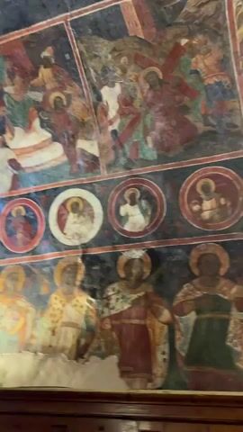 This video provides a close-up view of the interior of a church, focusing on its ornate ceiling and walls adorned with frescoes. The frescoes depict religious scenes and figures, including what appear to be saints or religious leaders in ornate attire, as well as smaller circular depictions of religious figures. The artwork shows signs of age and some damage, with areas of plaster missing or faded. A large, multi-tiered chandelier hangs from the ceiling, illuminated and adding to the grandeur of the space. The lower portion of the video shows wooden paneling, likely part of the church's structure or altar area. The camera pans and tilts, offering different perspectives of the detailed artwork and the overall atmosphere of the sacred space.