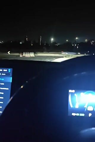 A person is driving a car at night. The car's dashboard touchscreen is visible, displaying the ambient light settings. The screen shows options to adjust the colors, brightness, and area of the ambient light, with a visual representation of the car. The car is driving on a road with streetlights and barriers visible in the distance. The car's windshield wipers are activated.
