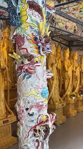 The video shows a detailed view inside a temple or shrine. In the foreground, a large, intricately decorated pillar is visible, covered in colorful mosaics depicting dragons and floral patterns. The pillar is white with vibrant accents of yellow, blue, red, and green. Behind the pillar, a row of golden statues stands arranged in a line. These statues appear to be Buddhist figures, possibly bodhisattvas, each standing upright with their hands in a prayer-like gesture. They are all golden in color and are placed on ornate golden pedestals. The background is adorned with colorful mosaic tiles, creating a rich and decorative environment. Small floral arrangements are placed at the base of some of the statues. The lighting suggests an indoor setting, possibly with artificial lights contributing to the bright illumination. The camera slowly pans from left to right, revealing more of the statues and the decorative surroundings. There are no people visible in the video.