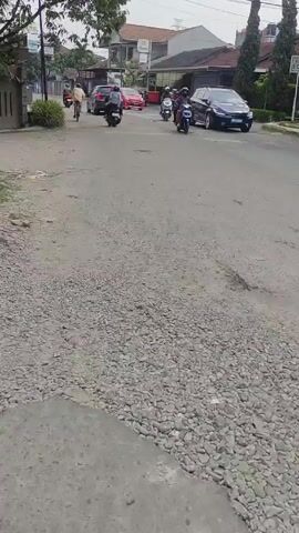 The video shows a street scene in a residential area. The road is paved but appears to be in disrepair, with patches of gravel and uneven surfaces. There are several vehicles moving along the street, including cars and motorcycles. A cyclist is also visible riding along the road. Buildings are visible on either side of the street, and there are trees and vegetation present. The overall lighting is overcast, suggesting a cloudy or dull day. The scene captures a typical moment of daily life in a suburban neighborhood.