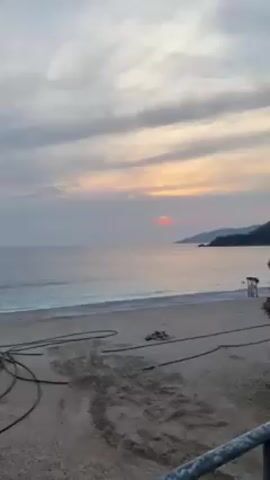 The video shows a wide shot of a beach at sunset. The sky is filled with clouds tinged with orange and pink from the setting sun, which peeks out just above the horizon over the ocean. A stretch of sandy beach is visible in the foreground, with a few dark cables or ropes lying on the sand. In the distance, there's a lifeguard stand and what appear to be forested hills along the coastline. The water is calm, with gentle waves lapping onto the shore. The overall scene has a serene and quiet feel.
