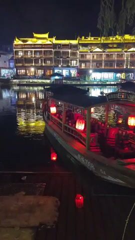 A video captures a boat at night, illuminated by red lanterns. The boat is docked by a wooden platform. In the background, a building with illuminated windows and traditional Chinese architecture stands tall. The water reflects the lights, creating a vibrant scene. The video appears to be taken from a slightly elevated perspective, looking down at the boat and the surrounding area.