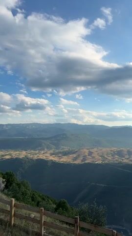 A video pans across a mountainous landscape under a partly cloudy sky. In the foreground, a wooden fence runs horizontally. The middle ground features rolling hills and a valley with a river winding through it. In the background, more mountains recede into the distance. The sun is visible through the clouds, casting rays of light onto the scene.