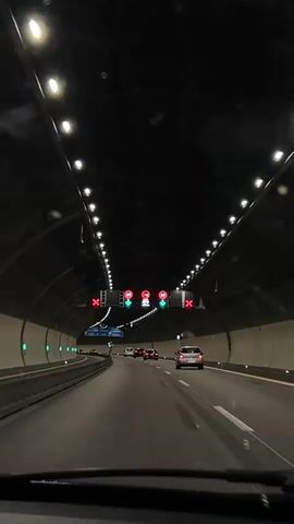 A camera is inside of a vehicle, driving on a road through a tunnel. Traffic in front of the vehicle includes multiple cars. The tunnel ceiling is dark, with rows of lights running along both sides. Speed limit signs with downward-facing green arrows above each lane indicate 60 kph is permitted for vehicles in the center lanes of the tunnel. Lanes on the left and right are closed, indicated by red X signs. The vehicle and other traffic appear to be in motion.