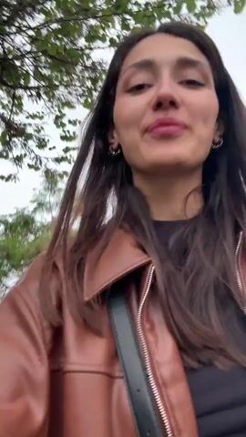 A young woman in a brown leather jacket is walking down a city street, holding the camera and recording herself. She smiles and then pauses for a moment as another woman walks up to her. The second woman is wearing a black leather jacket and holding a phone. They both smile at the camera as they stand in front of a building with the word "IDEA" on it. The two women then continue walking down the street together.