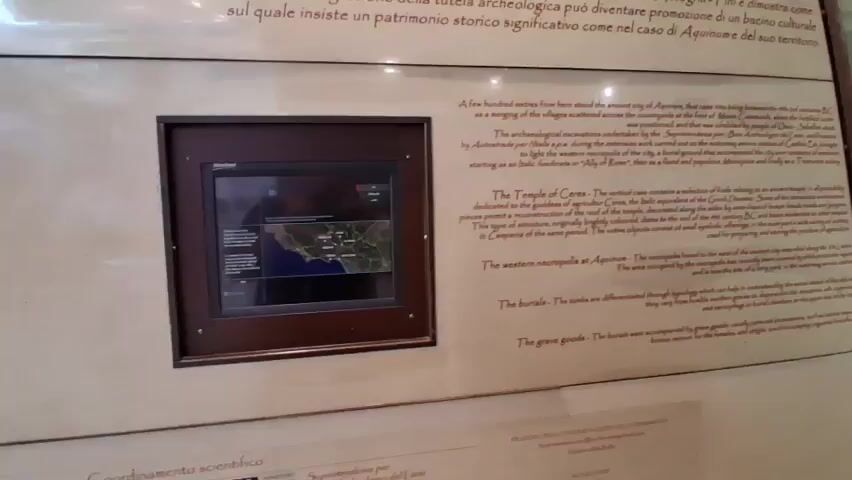 The video shows a close-up shot of a museum display case. The display case contains information about an ancient city. There is a screen on the left side of the display case that shows a map of an area. The text on the display case is in English and Italian. The text is about the ancient city of Aquinum, the Temple of Ceres, and the western necropolis at Aquinum. The video pans across the display case, showing more of the text and the screen.