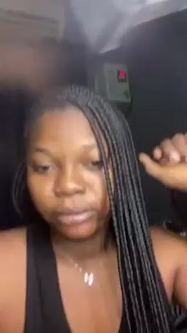 A woman is sitting in front of the camera. She has dark hair styled in braids, a black tank top, and a silver necklace with a bow pendant. She makes hand gestures and touches the pendant. An air conditioning unit and a device with a digital display are visible in the background. There is also a red light on the wall.