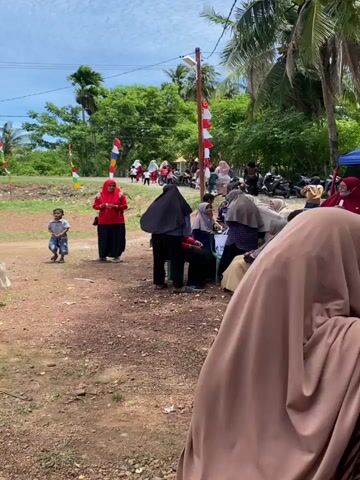 A group of people, mostly women wearing hijabs, are gathered outdoors on a dirt area. They appear to be participating in an event or celebration. Some individuals are seated under a tent, while others stand and mingle. A child is visible running in the foreground. In the background, there are trees, a cloudy sky, and what appears to be a road with a few motorcycles parked. Decorative flags, red and white, are displayed, suggesting a festive occasion.