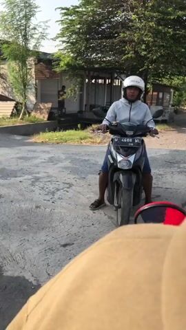 A person wearing a white helmet and a light-colored hoodie is sitting on a black motorcycle, looking towards the camera. They are wearing dark shorts. The motorcycle has a license plate that reads "4689 WL". The person's feet are on the ground. The motorcycle is parked on a paved surface with some cracked concrete. In the background, there is a building with a thatched or corrugated roof, and some trees. Another person is visible in the doorway of the building. The overall lighting suggests it is daytime, likely late afternoon. The camera appears to be positioned low, from the perspective of someone on another vehicle or sitting on the ground.