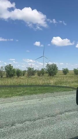 The video shows a view from a moving vehicle on a gravel road. In the background, a wind farm with several tall wind turbines is visible against a bright blue sky dotted with fluffy white clouds. The landscape is a mix of green and golden grasses with scattered trees. The scene appears to be set in a rural area, likely Kazakhstan, given the location context provided. The time of day appears to be daytime, judging by the bright sunlight and clear sky. The weather seems pleasant with a clear and sunny day. There are no people visible in the video. The focus is on the natural landscape and the industrial structures of the wind turbines. The overall mood is serene and open, suggesting a vast, undeveloped area. No visible text is present in the video.