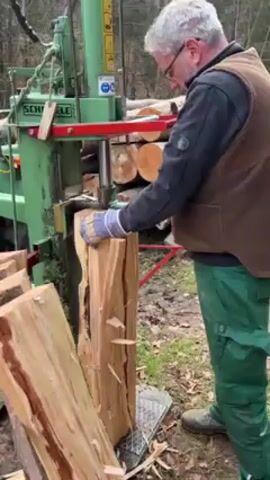 A man in green pants and a brown vest is operating a hydraulic log splitter. He’s wearing gloves. The log splitter is green and labeled “SCHIELE.” He's splitting a piece of wood that's already partially split.  Several other pieces of split wood are nearby. The environment appears to be wooded. The ground is covered in leaves and grass. There are a stack of unsplit logs in the background.