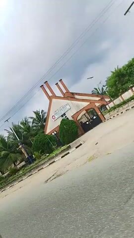 The video pans from right to left, showing the exterior of a building with a distinctive architectural design. The building has a peaked roof with multiple chimneys. In front of the building, there are palm trees and manicured bushes. A road runs in front of the building, with a sidewalk and a decorative border. The sky is overcast. There are no people visible in the video. The overall impression is of a well-maintained institutional or commercial building in a tropical setting.