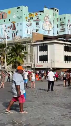The video shows a bustling public square or plaza during the day. Numerous people are walking around. Some are holding flags. There's a building with a colorful mural depicting people in traditional clothing. A palm tree is visible to the left. The sky is clear and blue.
