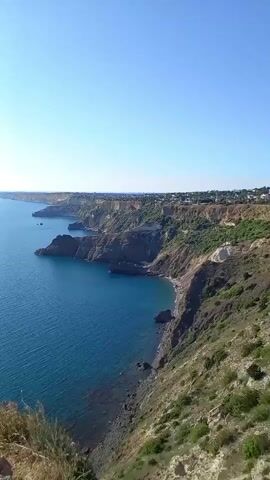 The video shows a panoramic view of a coastal landscape under a clear blue sky. The camera pans across a vast expanse of deep blue ocean meeting rugged, rocky cliffs dotted with greenery. In the distance, the coastline stretches out, revealing more cliffs and some scattered structures, suggesting a populated area or development along the shore. The sun is visible at one point, casting a bright glare on the water. The perspective is from a high vantage point, looking down and out towards the sea. There are no people or discernible objects in the water or on the land that would indicate specific activities. The environment appears to be natural and serene, with the focus on the grandeur of the ocean and the dramatic coastline.