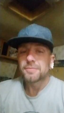 Okay, here is a detailed description of the video:

The video shows a man who appears to be in his late 30s to early 40s, based on his facial features. He has fair skin, light blue eyes, and a short, neatly trimmed beard. He also has a nose ring. His ears have been stretched and have white plugs in them. The man is wearing a light gray t-shirt and a blue baseball cap.

The man is facing the camera directly and speaking. He states he is reaching out to anybody that needs help or needs a friend and to contact him. 

The background is somewhat blurry, but appears to be the inside of a camper. There are cabinets visible above him and a patterned surface behind his right shoulder, suggesting some kind of furniture covering. The lighting is somewhat dim and overhead.

The video is static, meaning the camera is not moving. The quality of the video suggests it was recorded on a mobile phone.