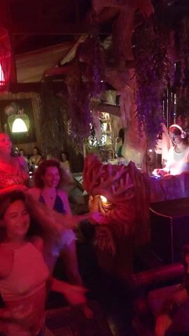 The video captures a lively nighttime party scene indoors, likely in a club or bar, at Lokaserena, Indonesia. The space is illuminated with vibrant artificial lighting, predominantly in shades of red and purple, creating an energetic atmosphere.

Numerous people, mostly young adults, are visible throughout the venue. They are engaged in various activities typical of a party, including dancing, socializing, and moving around the space. Some individuals can be seen closer to what appears to be a counter or bar area. A distinctive luminous white, fan-shaped decoration is briefly visible against a purple-lit background. The overall mood is festive and cheerful, with attendees appearing to enjoy the event. Music can be heard playing, consistent with a nightclub or party environment. No discernible text is visible within the video.