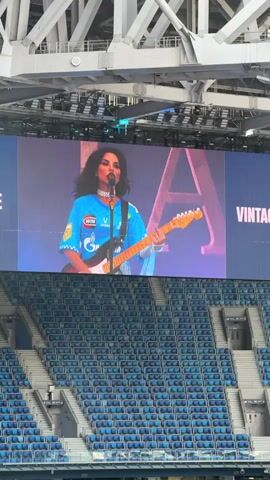 The video shows a large screen displaying a woman playing a guitar and singing. The screen is elevated above rows of empty blue stadium seats. The woman is wearing a blue shirt with logos and a necklace. The background of the screen has large letters, one of which is a prominent "A". To the left of the woman on the screen is another person playing a keyboard, and to the right, the word "VINTAGE" is visible. The stadium structure, made of white metal beams, is visible at the top of the frame.