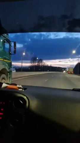 The video captures a view from inside a moving vehicle, likely a car, driving on a multi-lane road during dusk or early night. The sky above is a deep blue, with scattered darker clouds at the top of the frame, and a lighter, possibly purplish hue near the horizon. Streetlights illuminate the road ahead.

In the foreground, the dashboard and a portion of the steering wheel of the filming vehicle are visible. To the left, a large green dump truck with a light-colored, possibly beige, open bed is driving alongside the filming vehicle, then gradually moves ahead. Its red taillights are clearly visible. Further down the road on the right, another truck, appearing to be a semi-trailer, can be seen with its lights on.

The activity primarily involves driving on a highway or main road. Trees and sparse vegetation line the sides of the road in the distance. There is no visible text on any signs or vehicles that is clearly decipherable. The ambient sound includes music playing from a radio, featuring a male vocalist. The overall atmosphere suggests a routine evening drive. The location context provided is Beloborodovo, Russia, though no specific landmarks from this area are visible.