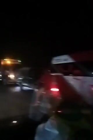 The video shows a night scene on a road with several vehicles. There are trucks and cars, all with their headlights on, creating streaks of light due to the camera's movement. The environment is dark, suggesting it is nighttime. Some vehicles have flashing lights, possibly indicating an incident or a service vehicle. The footage is blurry and shaky, suggesting it was captured from a moving vehicle. There are no clear views of people, but the presence of vehicles implies human activity. The scene appears to be a traffic situation, potentially a slowdown or blockage.