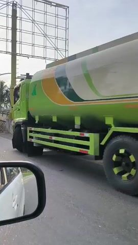 The video captures a side view of a bright green tanker truck driving on a road. The truck has a large cylindrical tank on its chassis, painted in shades of green and cream with curved designs. It has multiple axles and large black tires. Reflective red and white stripes are visible along the lower sides of the truck's chassis, likely for safety. In the foreground, a portion of a white car's side mirror is visible, indicating the video is shot from inside another vehicle. The truck is in motion, and the surrounding environment appears to be outdoors with a slightly overcast sky. A billboard structure on a tall yellow pole is partially visible in the background on the left, along with some trees and other road infrastructure. The truck is either passing or being passed by the vehicle from which the video is being recorded. The overall scene suggests movement along a public road.