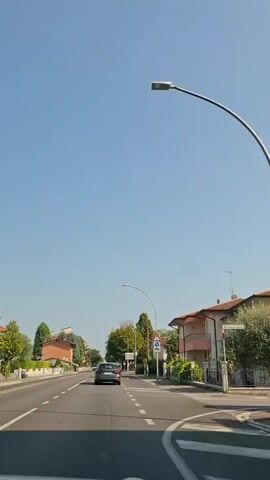 A vertical, eye-level shot shows a two-lane road with a clear blue sky overhead. A dark-colored car is traveling away from the camera in the right lane. White dashed lines mark the lanes, and white solid lines indicate the edges of the road. A white car is visible in the distance in the left lane. On the right side of the road, a traffic sign with a blue circular arrow indicating a mandatory turn to the right is visible, along with a red triangular warning sign. Houses with tiled roofs and lush green trees line the right side of the road. On the left side of the road, more houses and trees are visible. A lamppost curves over the road. The overall scene depicts a sunny day in a suburban or small town setting.