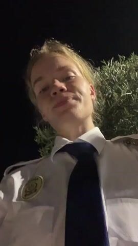 A person with blonde hair, wearing a white collared shirt and a dark tie, speaks into a camera. The person's face is illuminated by the camera's light. The background is dark with some blurred foliage visible. The person is close to the camera, and the footage is shaky.