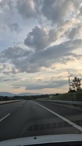 A point-of-view video from a car traveling down a highway, filmed from inside the car through the windshield. The road is clear with two lanes visible, separated by white lines. The sky is partially cloudy, and the sun appears to be setting, casting an orange glow on the horizon. Rolling green hills and fields flank the highway, with distant mountains visible on the left. A tall metal structure resembling a transmission tower stands on the right side of the road. Another vehicle is visible in the distance on the highway.
