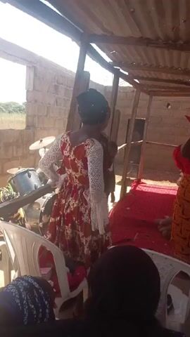 The video shows a gathering in an outdoor space, presumably for some type of ceremony. Several people are visible. A woman in a red top and patterned skirt stands near a small podium, apparently addressing the group. Other people, including children, are milling around or sitting in white plastic chairs. The area is covered by a corrugated metal roof, with brick walls forming the structure. A drum set is visible in the background, and there appears to be a red carpet on the floor of the platform. One man is taking photos or videos with his phone.