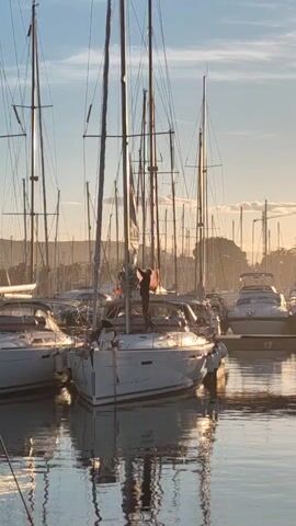 The video shows a marina with several sailboats moored at docks. The water is calm, reflecting the boats and sky. It's early morning, as the sun is low on the horizon, casting a golden light. A person is working on the mast of one of the sailboats. They are wearing dark clothing and appear to be making adjustments or repairs. Another person is standing on the deck of the same boat, possibly assisting. The scene is peaceful and indicative of a calm morning at a boat harbor.