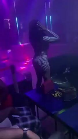 The video shows a woman dancing in a nightclub. She's wearing a short, striped dress, and a bright pink purse sits on a table next to her. The club is dimly lit with colorful, flashing lights and smoke effects. A speaker and a couch are visible in the background. The camera focuses on the woman's movements as she dances to the music.