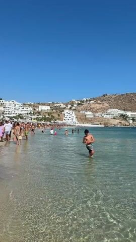 The video shows a clear blue sky over a beach with a hillside covered in white buildings in the background. The water is clear and turquoise, with gentle waves. Several people are visible on the beach and in the water. In the foreground, a person in patterned swim trunks stands in the shallow water, looking down at their hands. Further out in the water, another person is swimming. Along the shoreline, a crowd of people are walking, standing, and swimming. Several boats are anchored in the distance. The scene appears to be a popular tourist destination on a sunny day.
