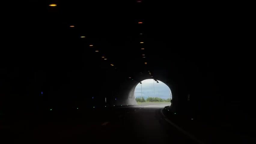 The video shows a car driving through a dark tunnel, gradually approaching the exit. The tunnel is lit by dim lights, and the road inside is clear. As the car gets closer to the exit, the outside world becomes visible, revealing a cloudy sky, a road with streetlights, and some greenery. The road outside the tunnel curves slightly. The car continues to drive out of the tunnel, transitioning from the dark interior to the overcast exterior. The video appears to be shot from inside the vehicle. Electronic dance music is playing in the background.