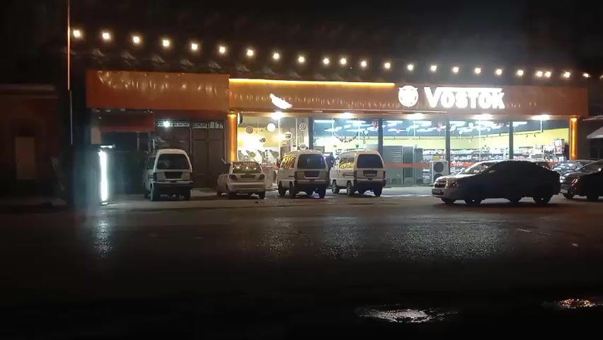 The scene is set at night, in front of a store called "VOSTOK", which is illuminated by a string of lights. Several cars are parked in front of the store, including a few white vans. The street appears wet. A few people are visible in the distance.