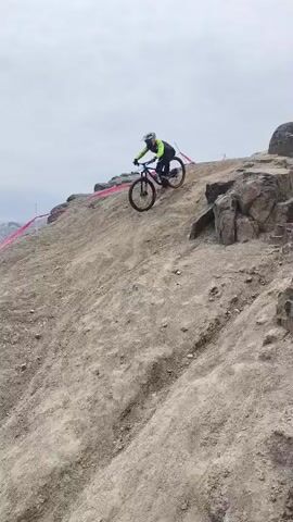 A mountain biker, wearing a neon yellow/green long-sleeved jacket, black pants, and a full-face helmet, is captured descending a steep, rocky, and dusty hillside. The individual is riding a black full-suspension mountain bike, skillfully navigating the challenging, uneven terrain.

The scene is set on an elevated, arid landscape overlooking a sprawling cityscape under an overcast or hazy sky. In the background, numerous buildings and roads of a city stretch out towards distant mountains. Red and white tape is visible along parts of the trail, suggesting a marked course for an event or race.

As the biker continues their descent, more details of the environment emerge. A large, light-colored building, possibly industrial, and a distinctive circular structure with a blue rim are visible among the urban sprawl. Further down the slope, several other individuals are gathered, some with bikes, appearing to watch the biker or participate in the event. Cars and motorcycles are also parked in the background. The ground is dry, loose, and covered with rocks and earth. The overall impression is an outdoor mountain biking activity taking place on a rugged, elevated trail in Trujillo, Peru. No legible text is visible on any of the subjects or objects.