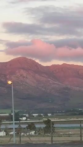 The video depicts a view of a mountain range, tinted pink by the setting sun. The sky is a mix of pale pink, light blue, and grey clouds. In the foreground, there's a landscape of trees, buildings, and fields. Various objects, like a streetlight and roadside signs, are seen as the camera appears to be moving past in a vehicle.