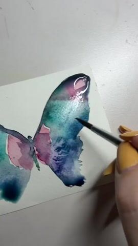 A close-up shot focuses on a hand painting a watercolor butterfly. The butterfly's wings are a blend of blues, greens, and purples, with a touch of pink. A paintbrush, held by a person with yellow-painted fingernails, is actively applying paint to the edge of the butterfly's wing. The paper the butterfly is painted on is white and textured. The background is a plain, light-colored surface, likely a table or desk. The lighting is bright and focused on the artwork.